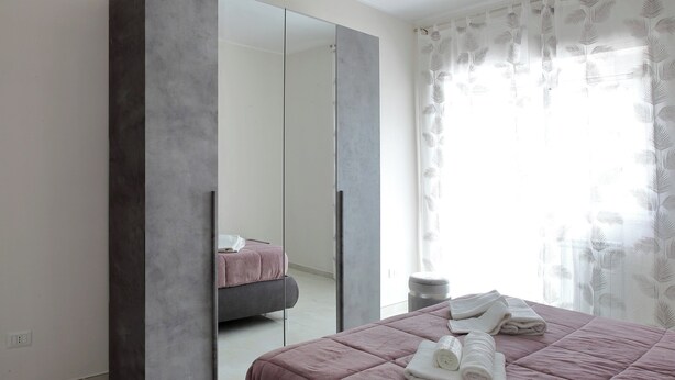 Una camera da letto con un grande specchio, un letto con lenzuola rosa, un piccolo sgabello e una finestra con tende leggere.