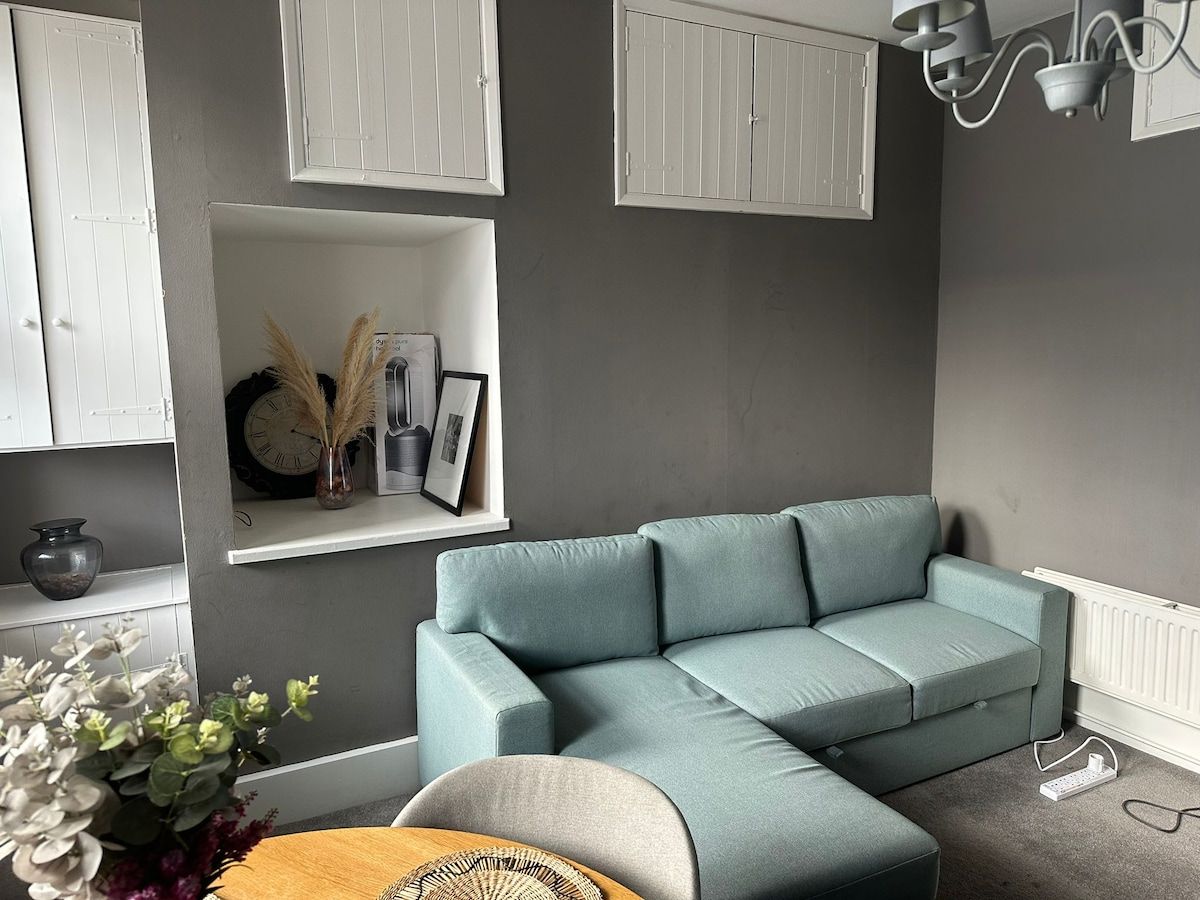A living room with a light blue sectional sofa, a small round table with a vase of flowers, and a white cabinet with a clock and picture frames.