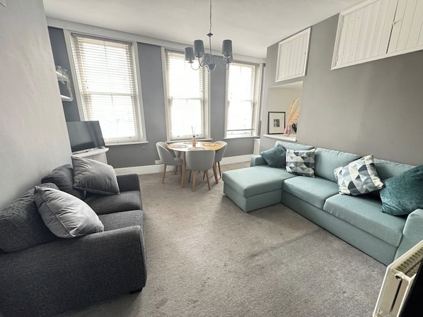 A modern living room with grey sofas, a round dining table, and a chandelier.