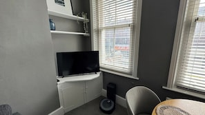 A room with a flat-screen TV on a white cabinet, a small speaker, a round table with a woven basket, and a window with blinds.
