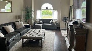 A living room with a black leather sofa, a grey sofa, a tufted ottoman, a wooden cabinet, and a large flat-screen TV. - Myrtle Beach Condo in Kingston Property steps away from Ocean, pool, food & fun (Myrtle Beach)
