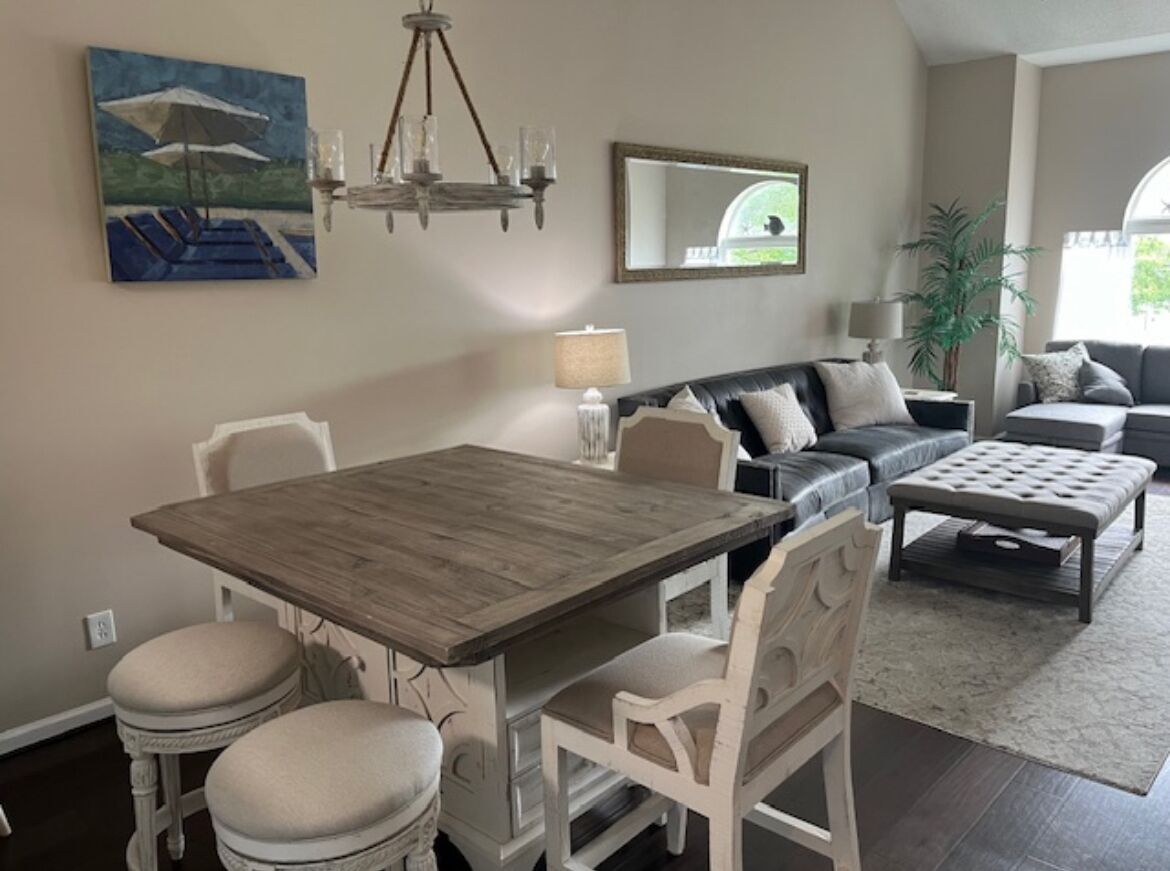 Een eethoek met een houten tafel en witte stoelen, een bank met kussens, een lamp en een schilderij aan de muur.