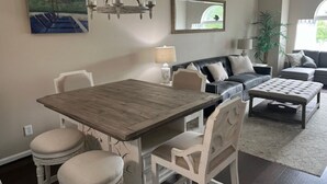 A dining area with a wooden table and white chairs, a sofa with cushions, a lamp, and a painting on the wall. - Myrtle Beach Condo in Kingston Property steps away from Ocean, pool, food & fun (Myrtle Beach)