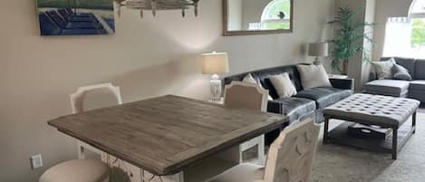 Een eethoek met een houten tafel en witte stoelen, een bank met kussens, een lamp en een schilderij aan de muur.