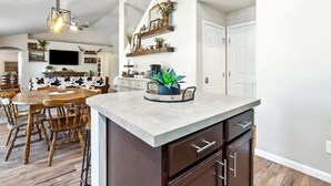 Una cocina moderna con una isla central, una mesa de comedor de madera y sillas. Cuenta con un horno empotrado y un ventanal que proporciona luz natural.