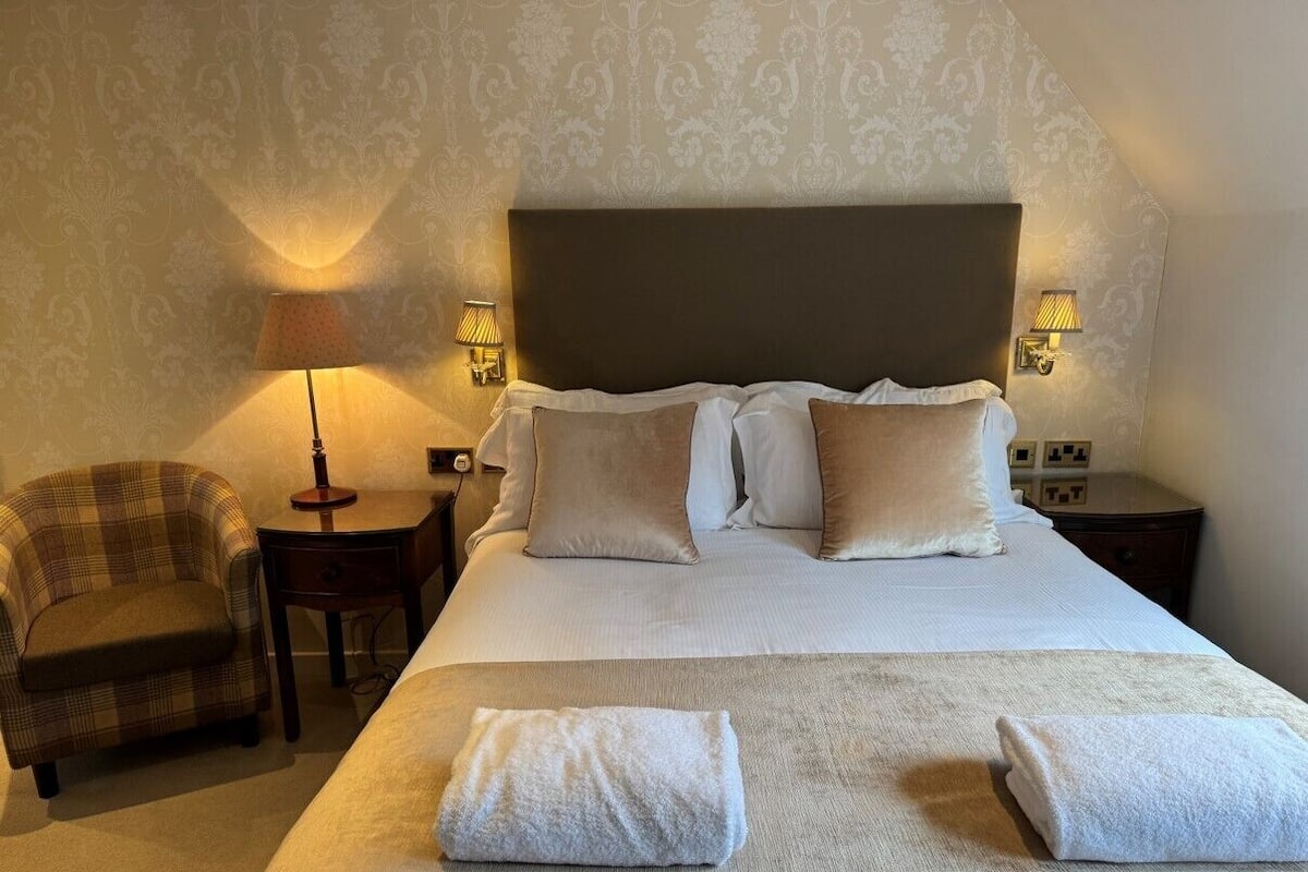Una habitación de hotel con una cama grande, dos lámparas de noche, una silla y una mesita.