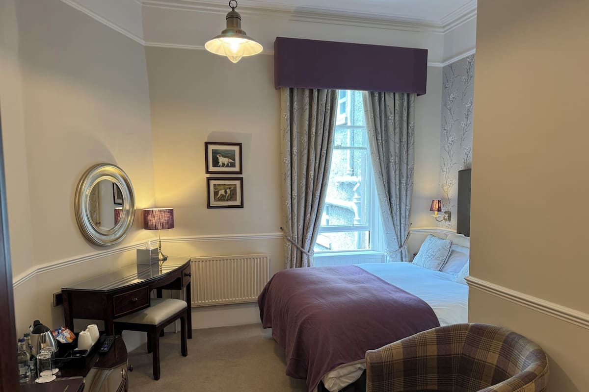 Una habitación de hotel con una cama, un escritorio con lámpara, una silla, una ventana con cortinas y un espejo montado en la pared.