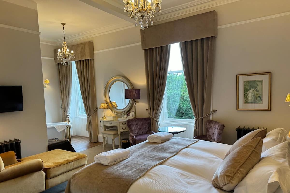 Habitación de hotel con una cama grande, dos sillones, un tocador con espejo, una araña y una ventana con cortinas.