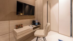 A modern hotel room with a flat-screen TV mounted on the wall, a wooden vanity with a mirror, a small shelf with toiletries, and a cushioned chair.