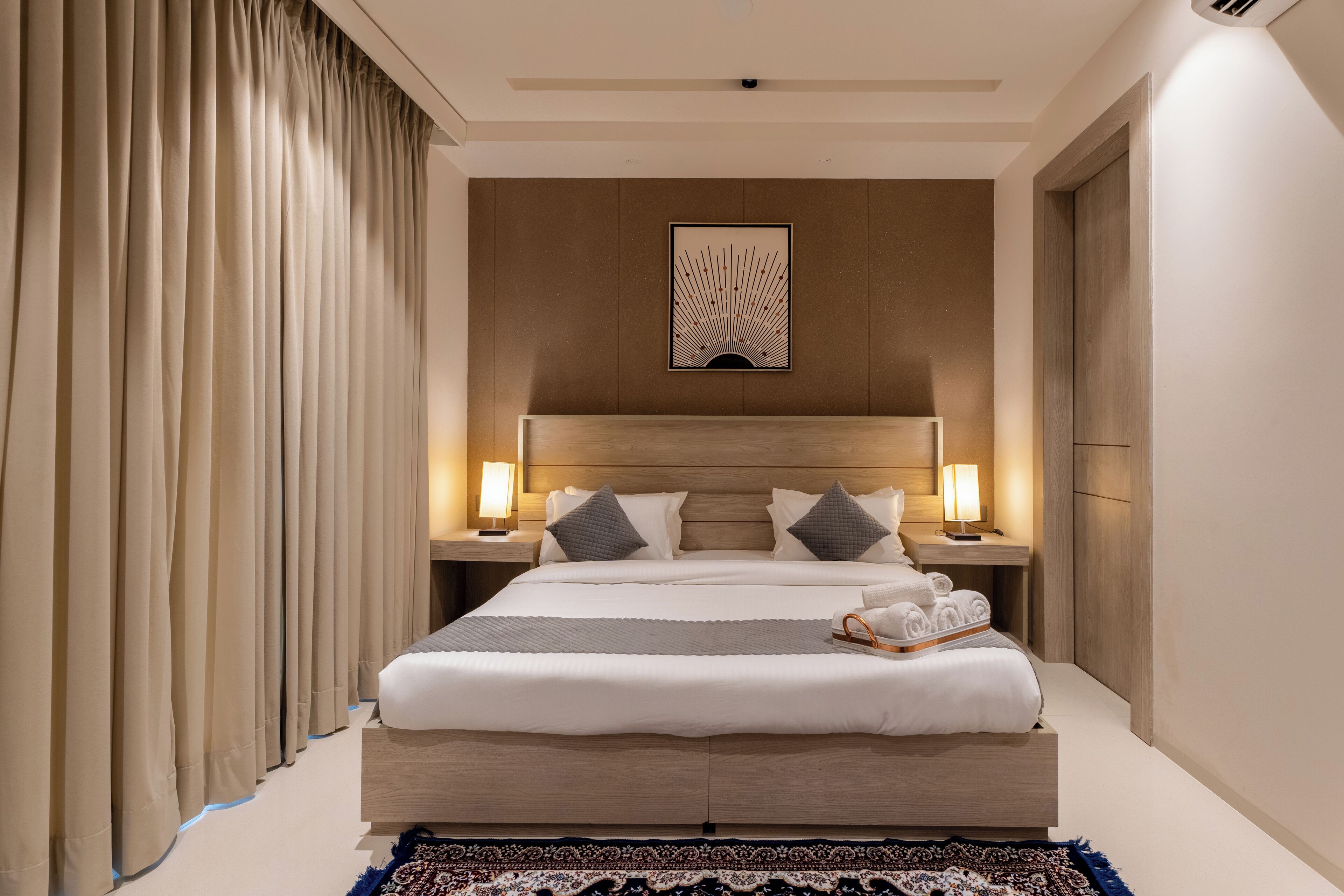 A hotel room with a large bed, two bedside tables with lamps, a headboard with a decorative panel, and a large window with curtains.