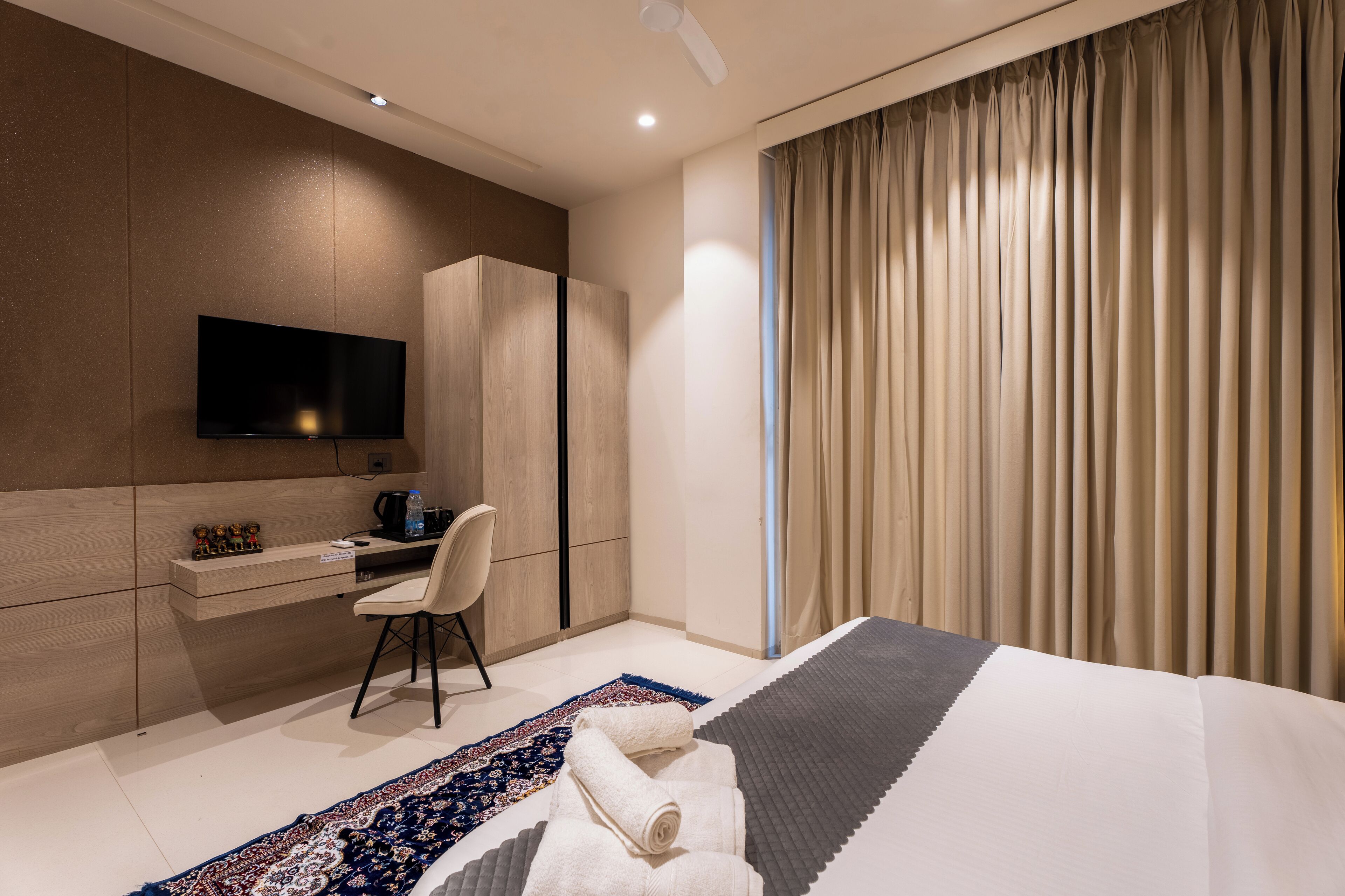 A modern hotel room with a flat-screen TV, a desk, a chair, a bed with a patterned rug, and a large window with curtains.