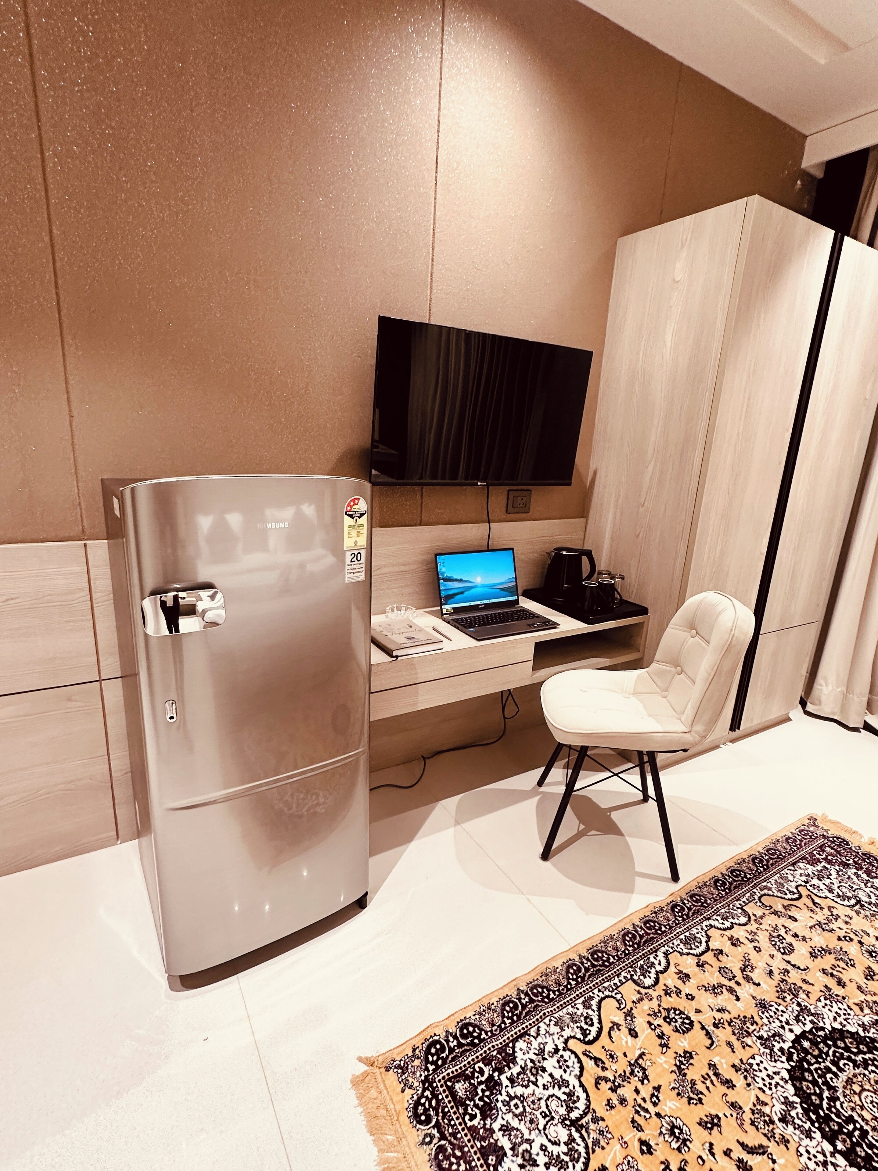 A modern kitchen with a stainless steel refrigerator, a laptop on a wooden countertop, a white chair, and a flat-screen TV mounted on the wall.