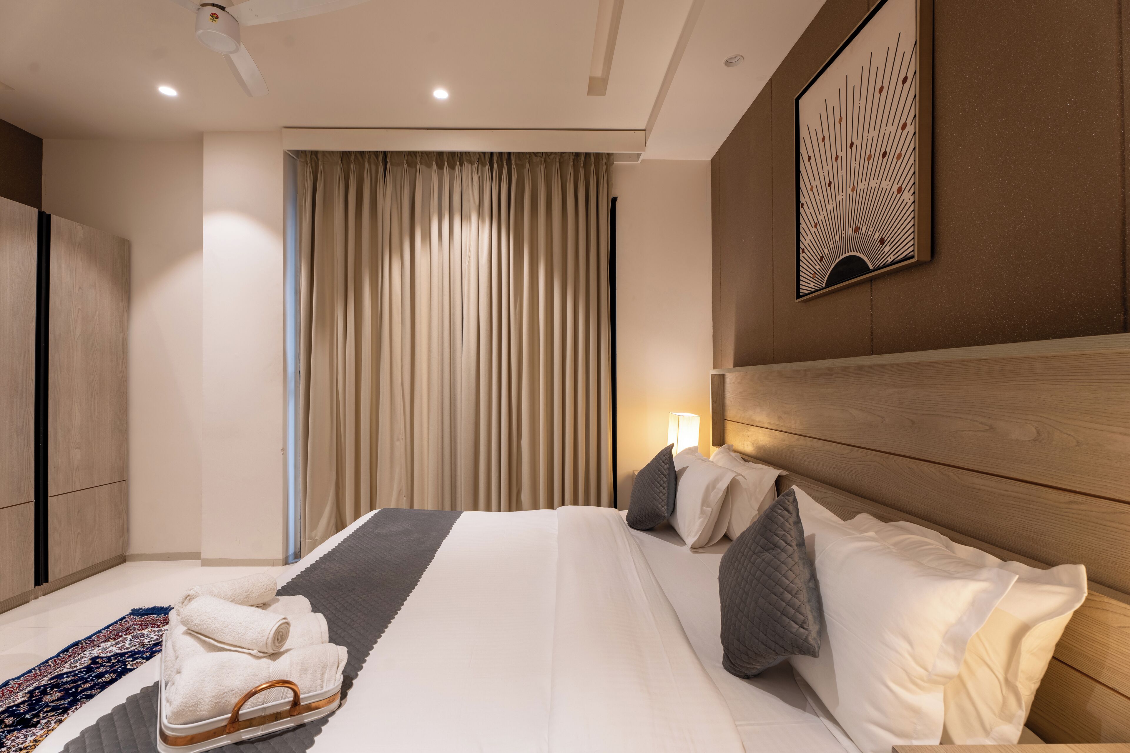 A modern hotel room with a large bed, a wooden headboard, a bedside table with a lamp, and a view of the city through floor-to-ceiling windows.
