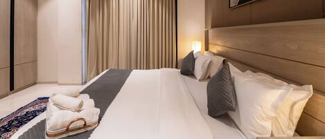 A modern hotel room with a large bed, a wooden headboard, a bedside table with a lamp, and a view of the city through floor-to-ceiling windows.