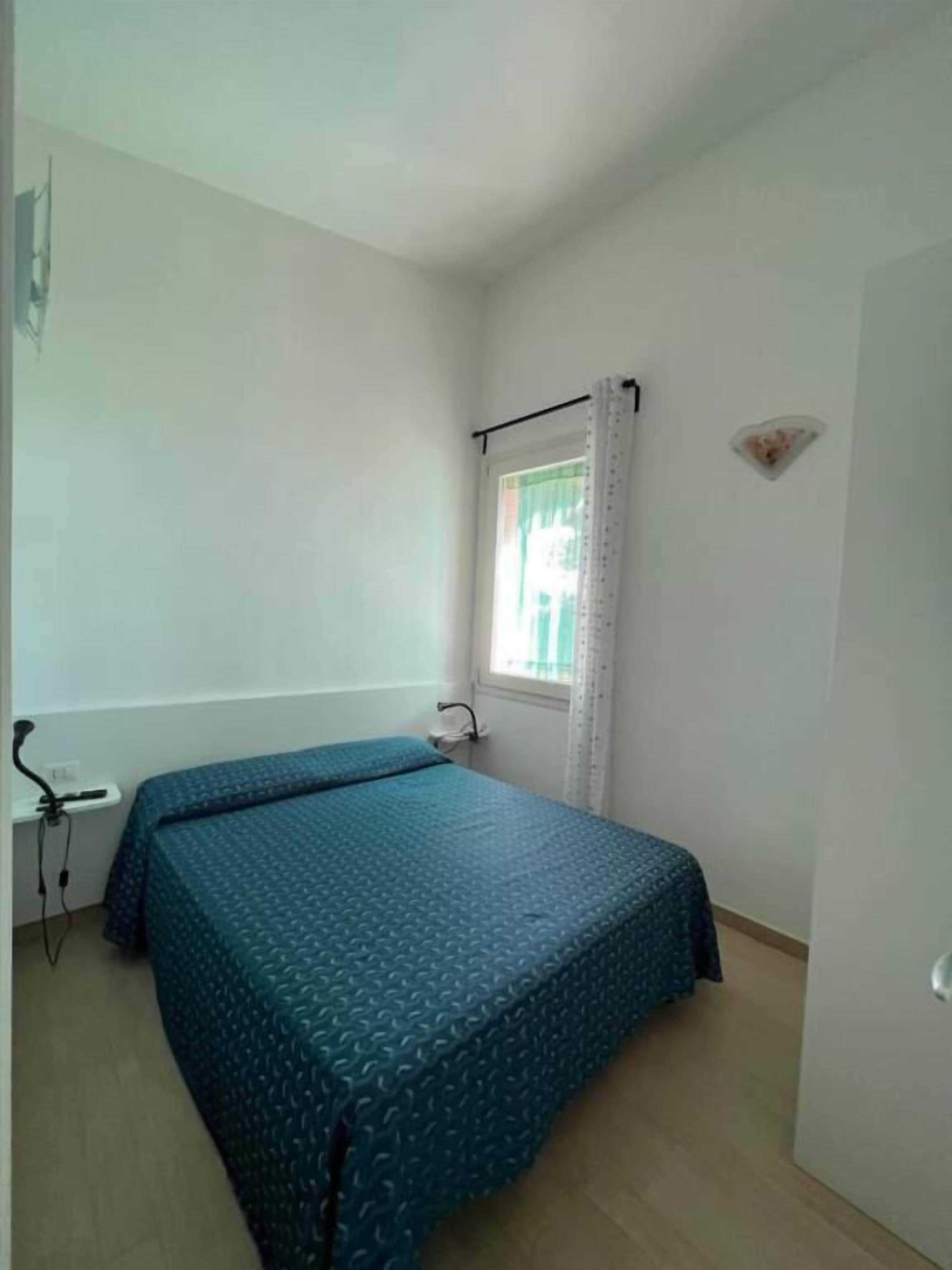 Habitació petita i poc moblada, amb un llit sol, una finestra amb cortines i una porta.