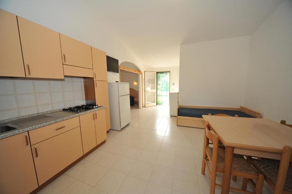 A compact kitchen with beige cabinets, a stove, and a refrigerator. There is a wooden dining table and bench in the adjacent dining area.