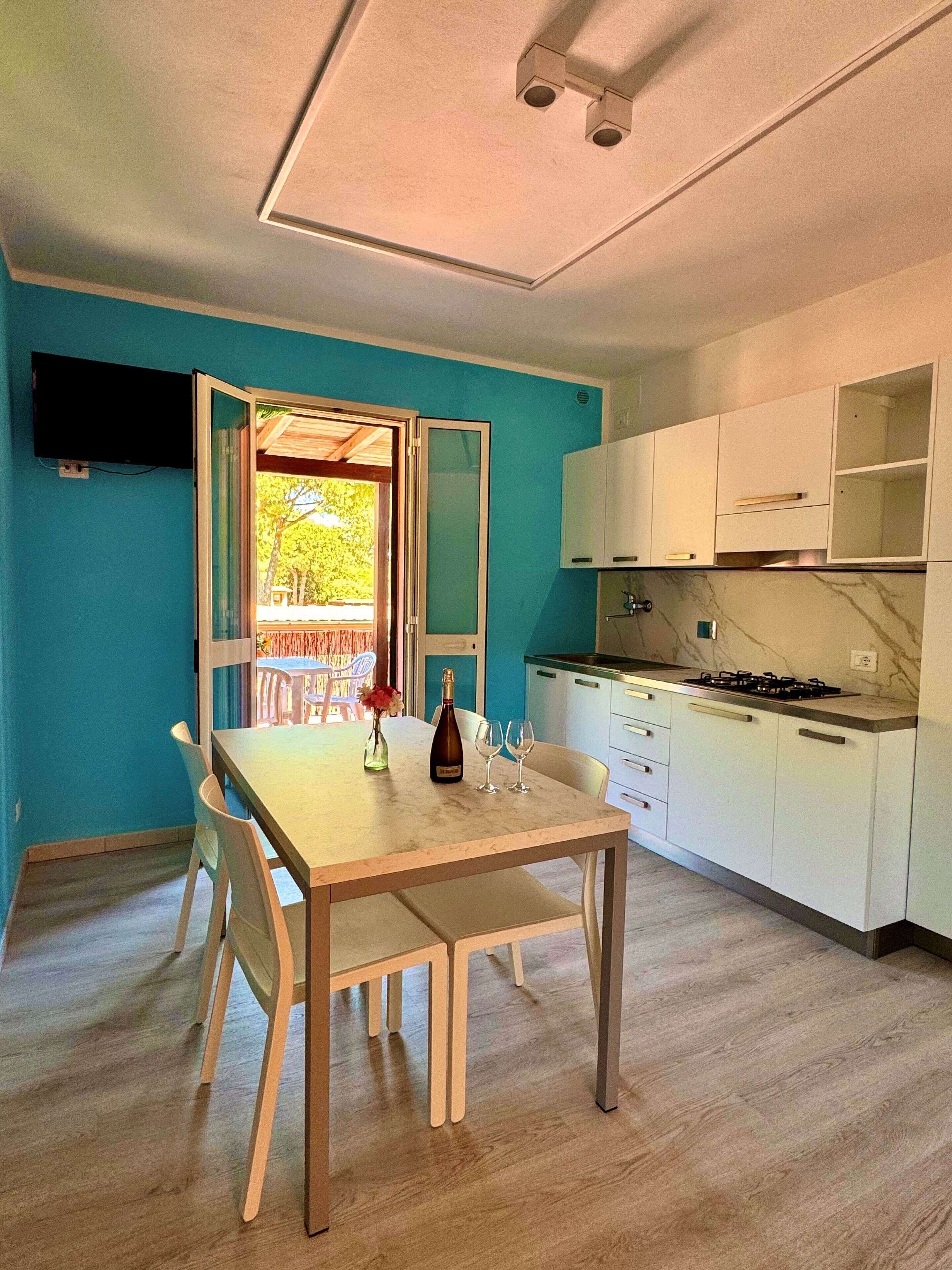 Una cucina compatta con mobili bianchi, una parete di fondo in marmo e un'area pranzo con tavolo e sedie in legno.