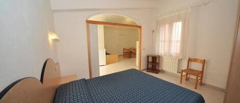 Un dormitorio con una cama, un escritorio, una silla y una mesita. Hay una puerta que da a otra habitación.