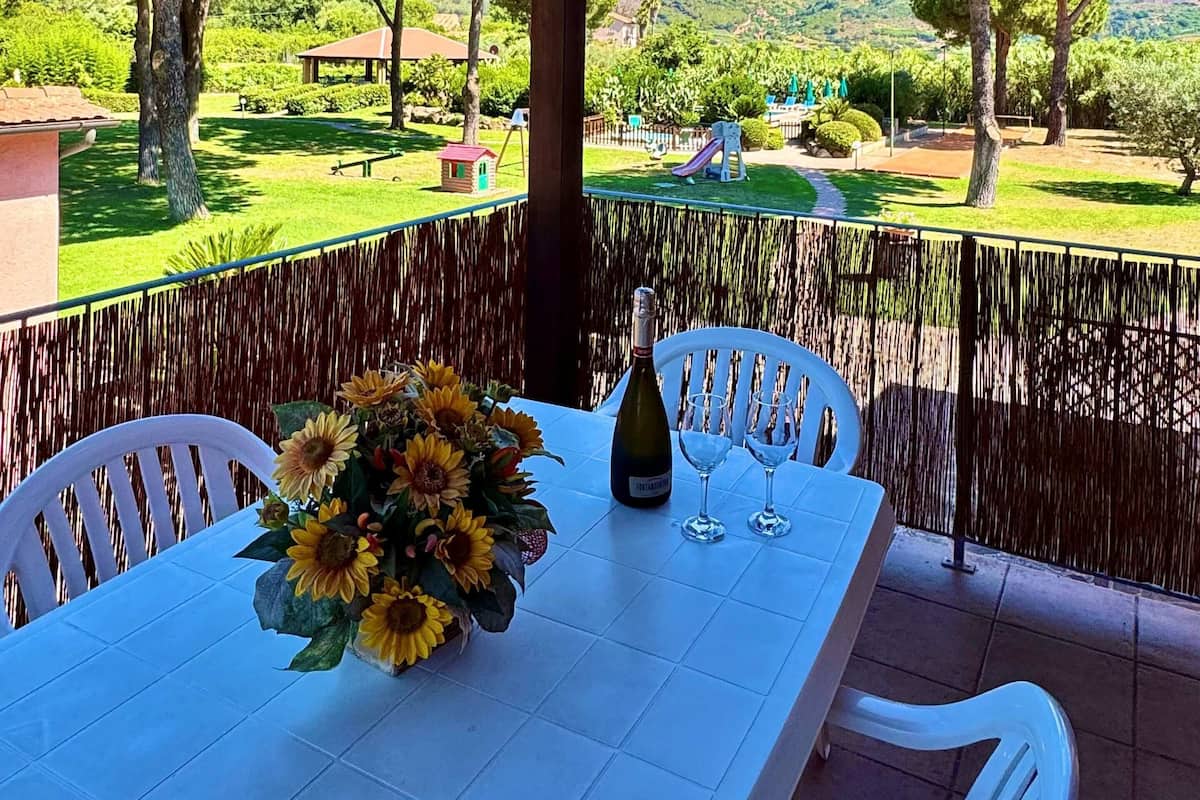 A covered outdoor dining area with a table set for two, a bottle of wine, and a vase of sunflowers.