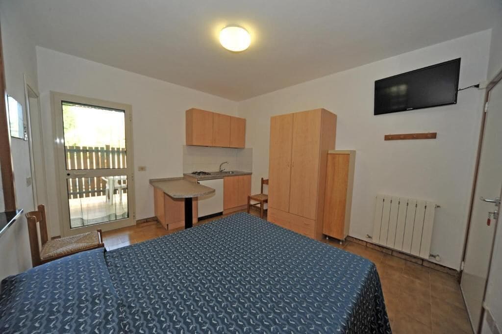 Une pièce compacte comprenant un lit, une petite kitchenette, un téléviseur et une porte coulissante donnant sur un espace extérieur.