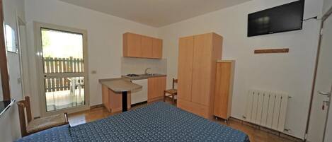 Una habitación compacta con cama, una pequeña kitchenette, televisor y puerta corrediza a un área exterior.