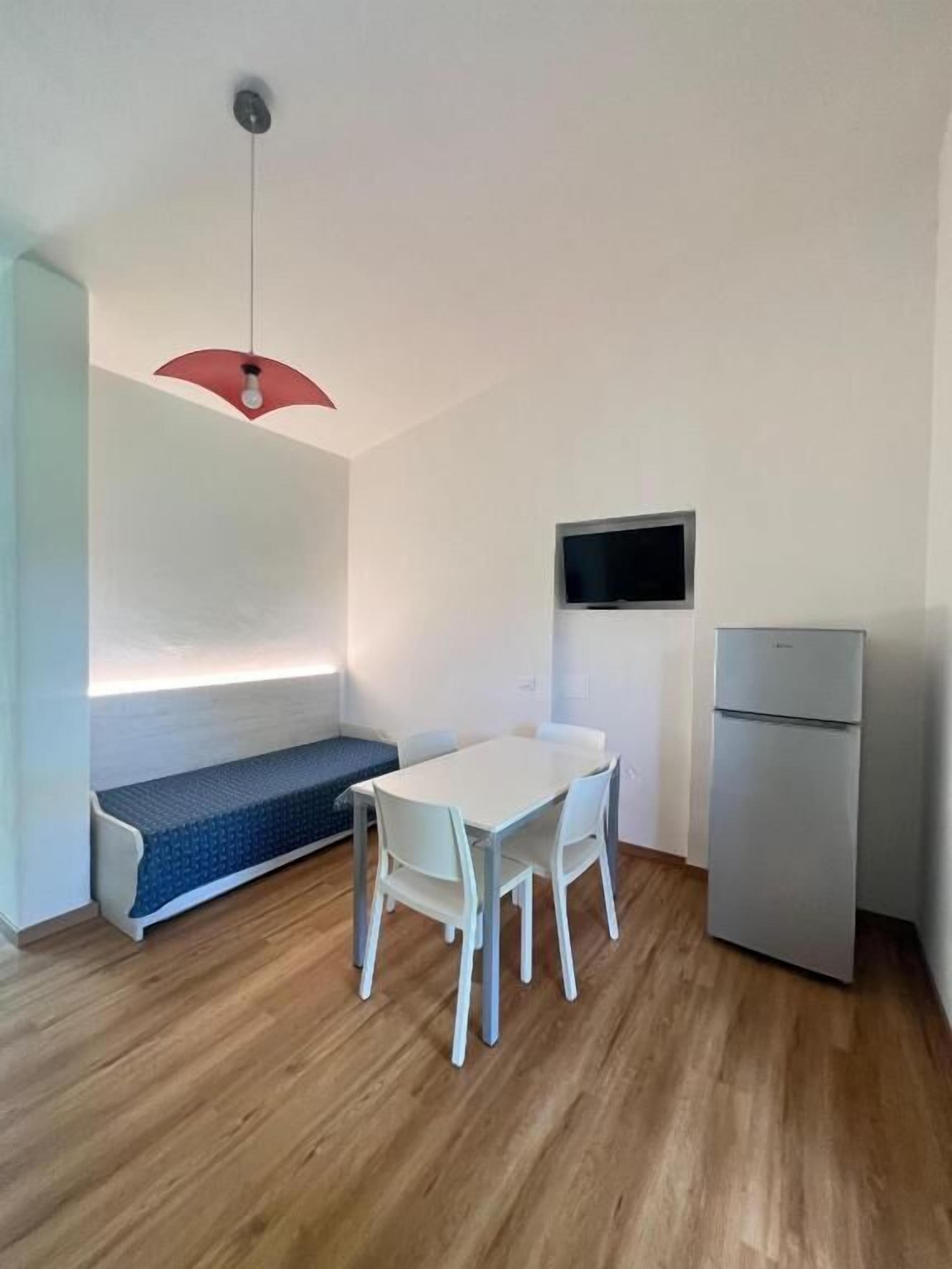 A small, modern kitchen with a dining table, chairs, a bench, a refrigerator, and a TV mounted on the wall.