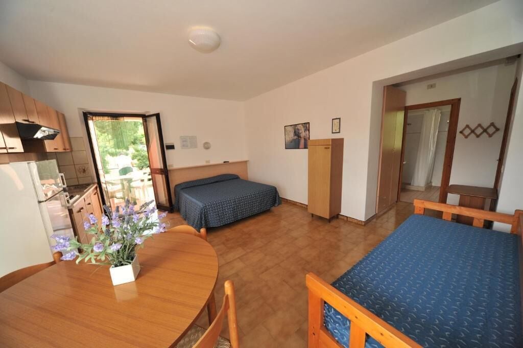 A compact room with a wooden dining table, a single bed, a small kitchenette, and a door leading to an outdoor area.