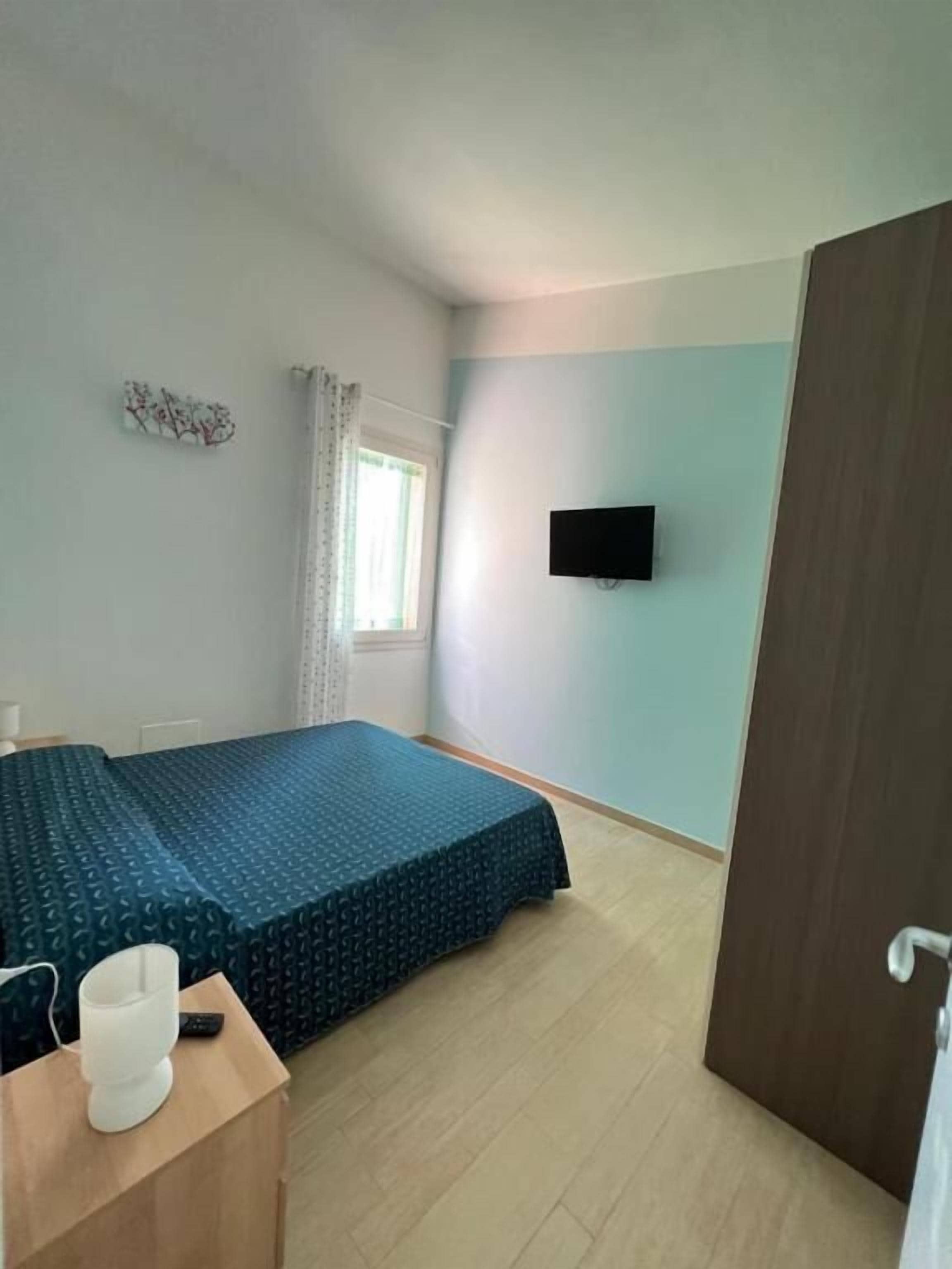 Une pièce comprenant un lit, un téléviseur fixé au mur, une fenêtre avec des rideaux et une porte en bois.