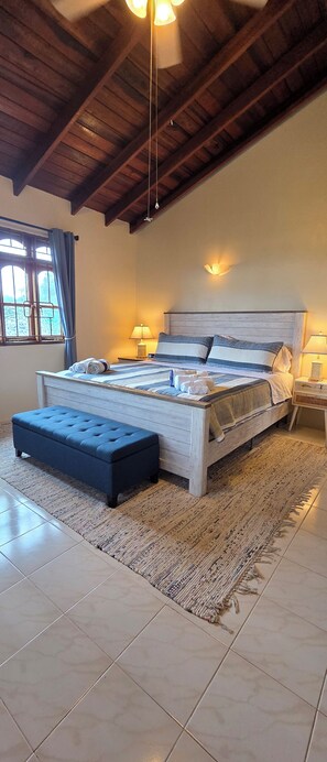 A bedroom with a large bed, a nightstand, a lamp, a window with curtains, and a ceiling fan. - Spacious Platinum Coast ranch villa with pool & AC close to the beach (Heywoods)