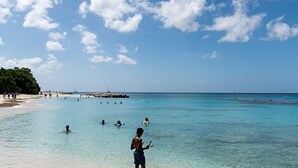 A beach with clear turquoise water, people swimming and fishing, and a rocky outcrop in the distance.