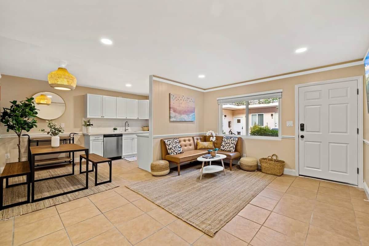 A spacious living area with a sofa, coffee table, and dining table. There is a kitchen with white cabinets and a dining area with a plant and a mirror.