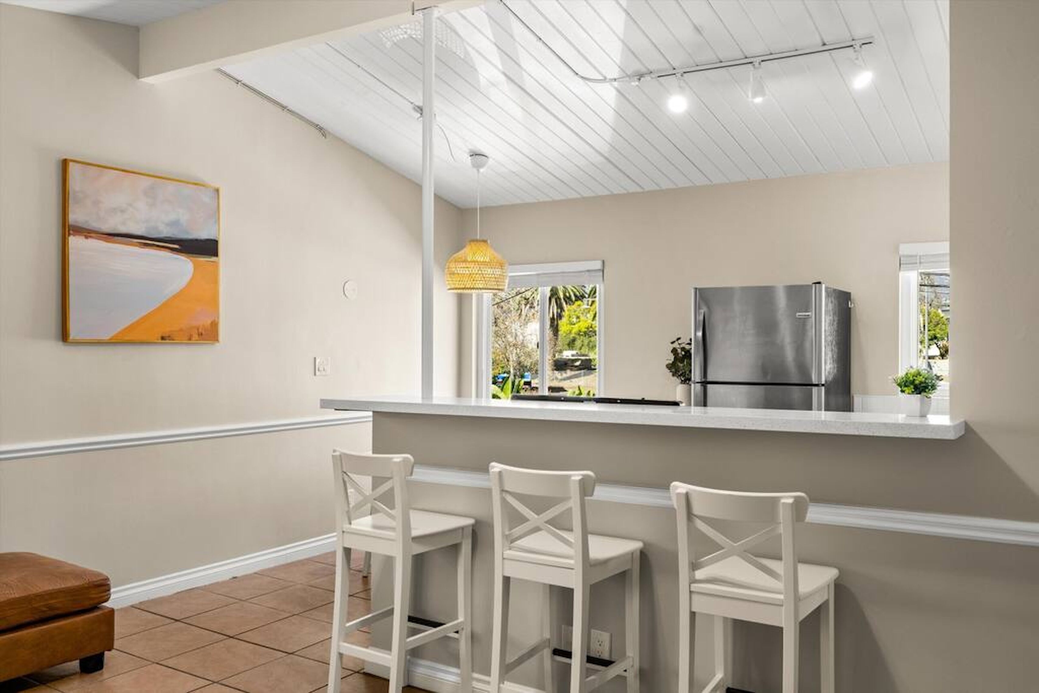 A modern kitchen with a white countertop, three white bar stools, a stainless steel refrigerator, and a hanging pendant light.