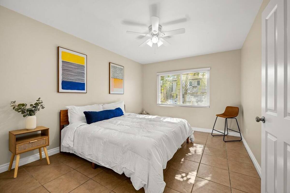 A bedroom with a bed, a ceiling fan, a window with blinds, a chair, and two framed artworks on the wall.