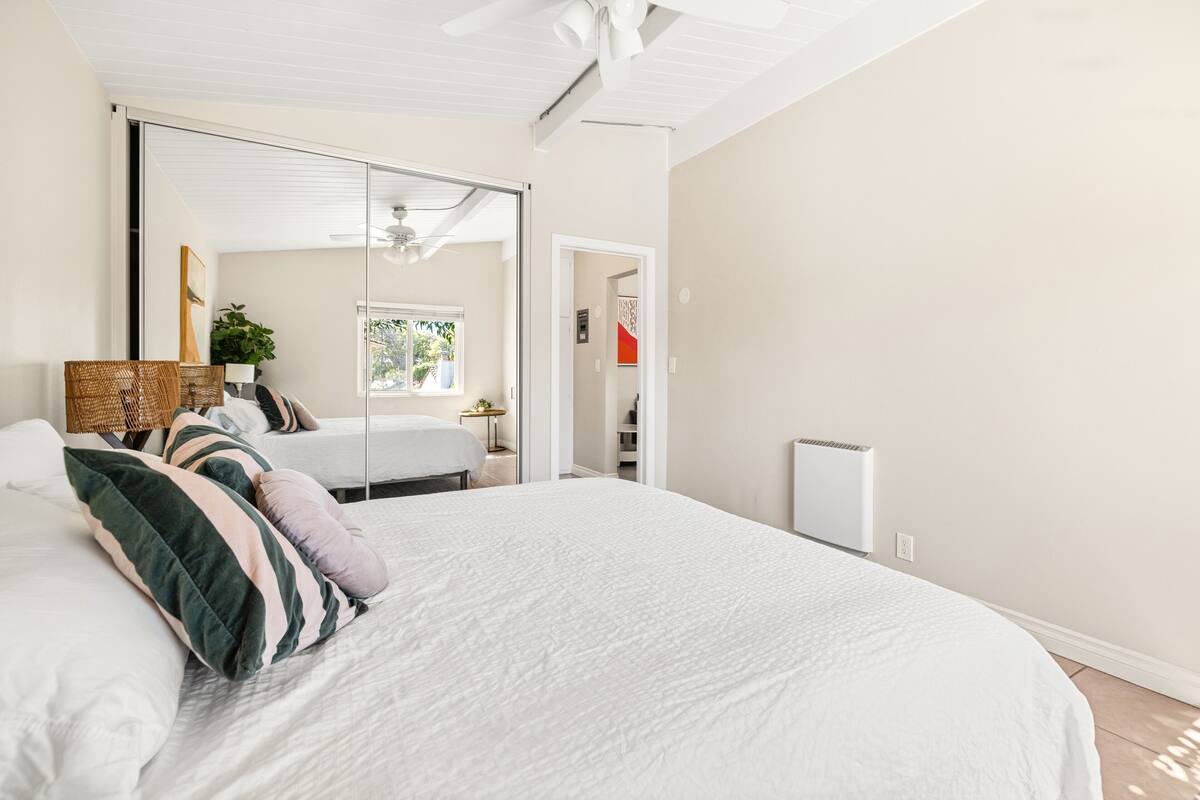 A bedroom with a large bed, a ceiling fan, a sliding door leading to a bathroom, and a small table with a plant.