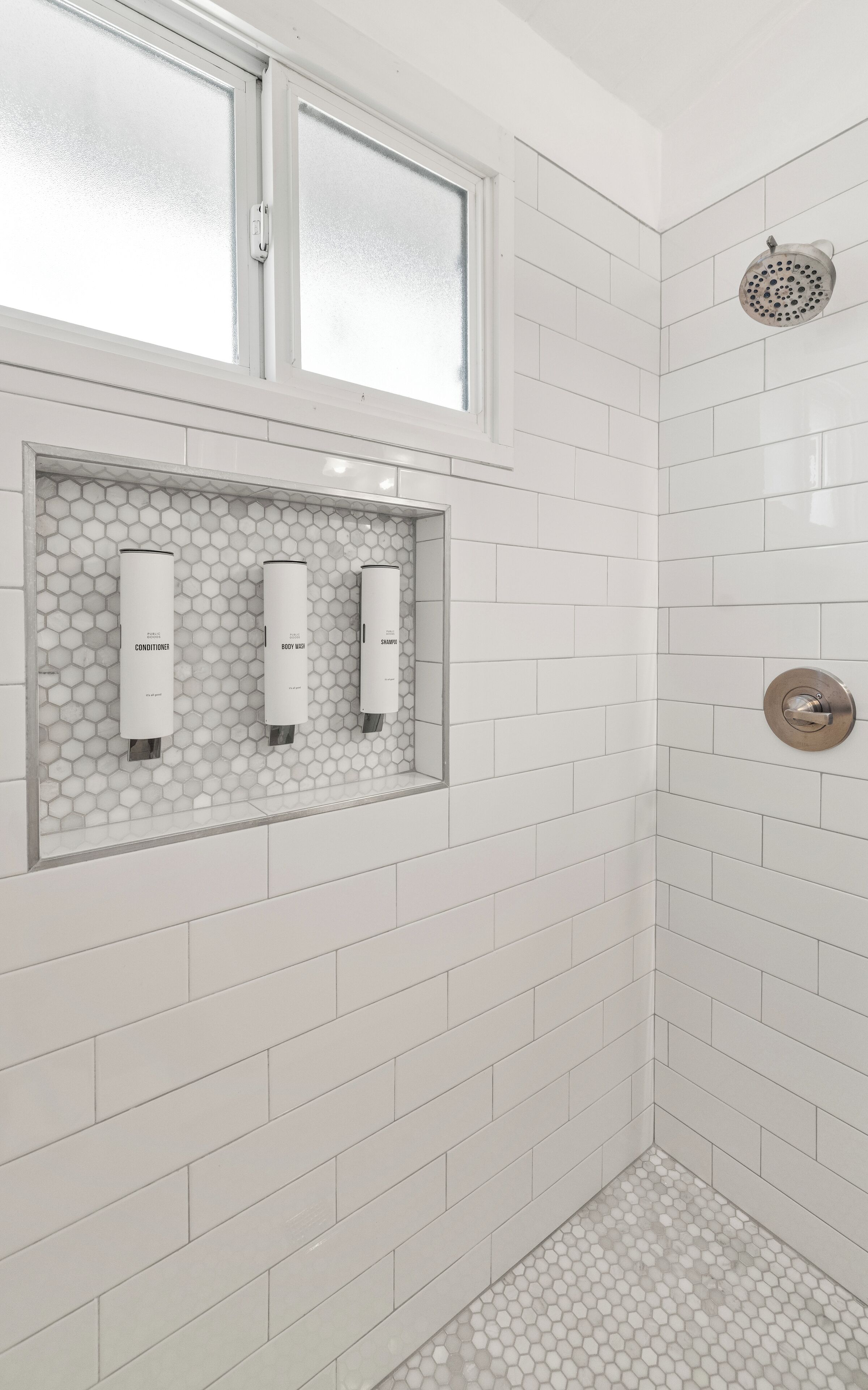 Une salle de bain avec des carreaux blancs, une douche dotée d’un pommeau de douche à effet pluie et deux distributeurs de savon fixés sur un mur recouvert de carreaux hexagonaux.