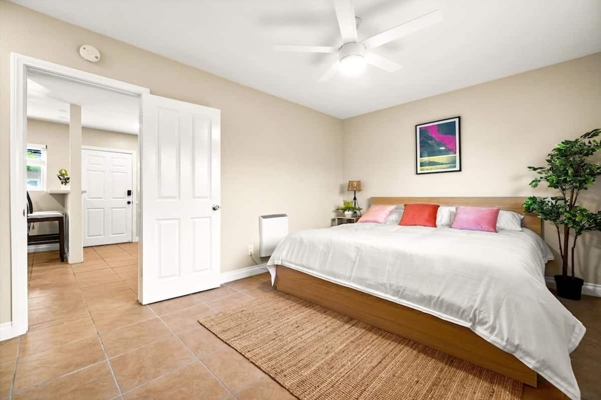 A bedroom with a bed, a ceiling fan, a nightstand, a lamp, a plant, and a door leading to another room.