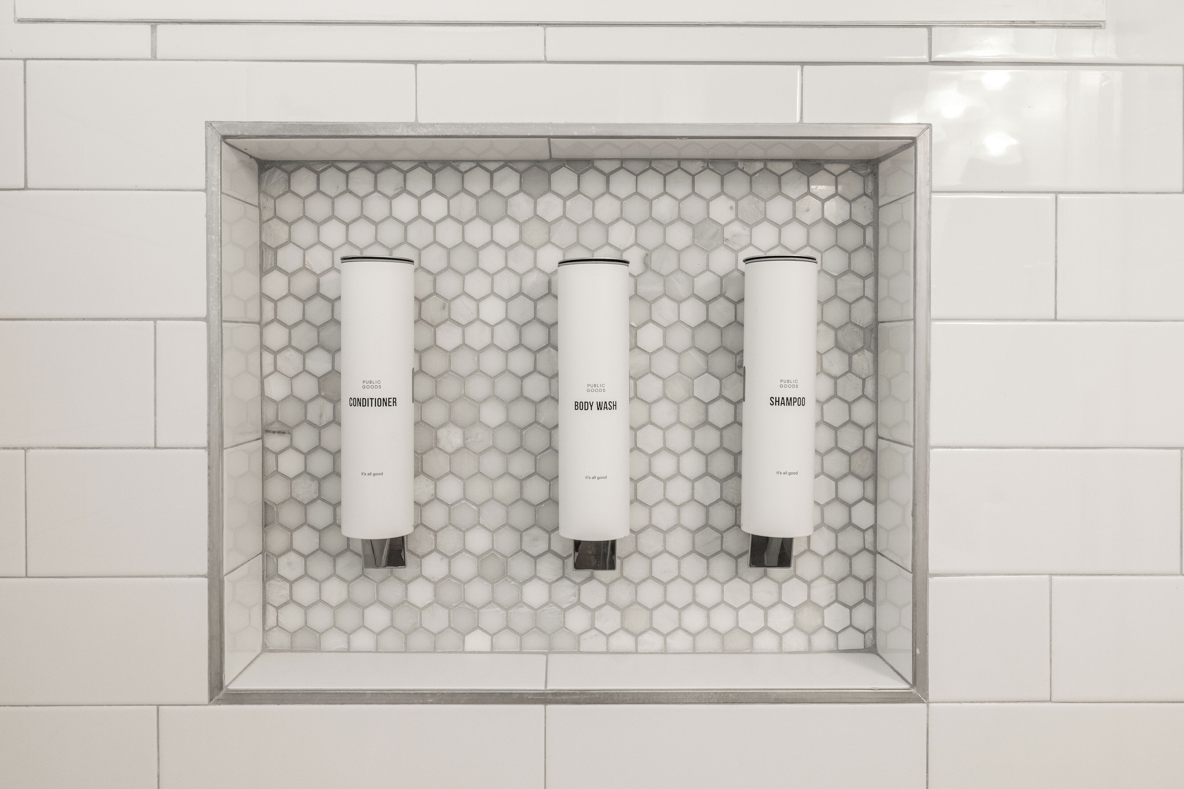 Une salle de bain avec trois flacons blancs étiquetés « Après-shampoing », « Gel douche » et « Shampoing », le tout sur un mur recouvert de carreaux hexagonaux.