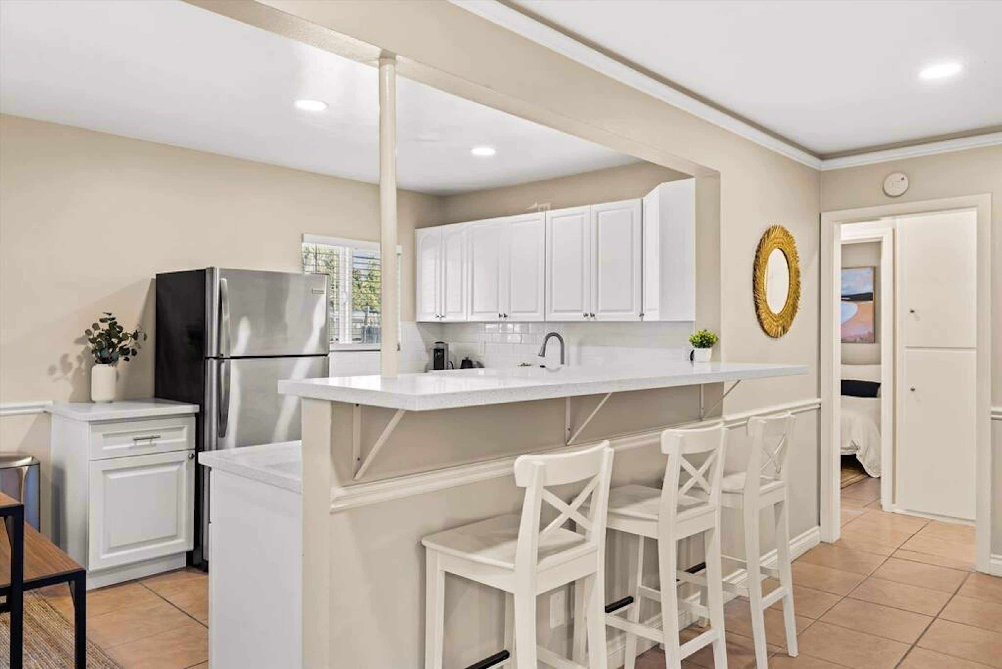 A modern kitchen with white cabinetry, a stainless steel refrigerator, a white island with bar stools, and a decorative gold wreath on the wall.