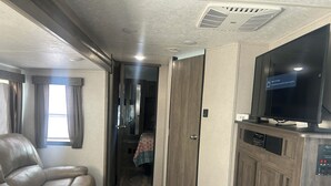 Un pasillo estrecho con un sillón de cuero, un televisor sobre un mueble, y una puerta que da a un dormitorio.