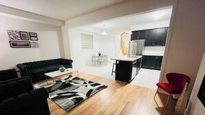 A modern living room with a black sofa, a coffee table, and a patterned rug. There is a kitchen area with dark cabinets and a red chair. - Spacious & Modern 4 BR House (Brantford)