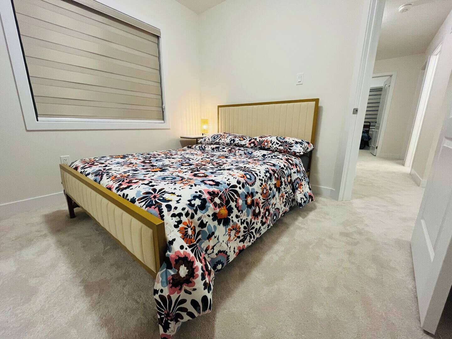 Ein Schlafzimmer mit einem Bett, einem Nachttisch, einem Fenster mit Jalousien und einer Tür zu einem anderen Raum.