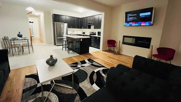 A modern living room with a black sofa, a white coffee table, and a patterned rug. There is a kitchen area with dark cabinets and stainless steel appliances. A flat-screen TV is mounted on the wall, and a red chair is visible in the corner. - Spacious & Modern 4 BR House (Brantford)