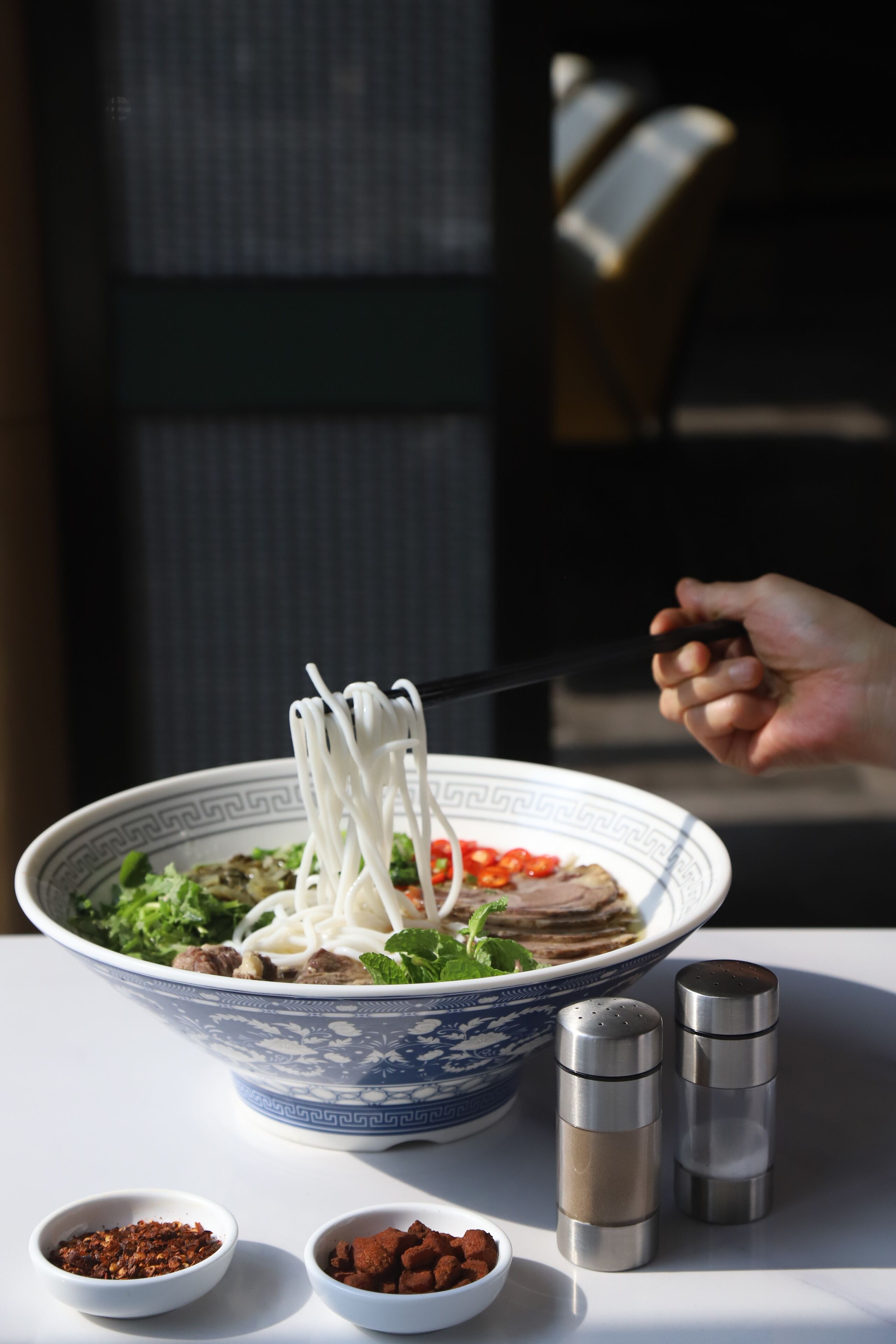 A bowl of noodle soup with various toppings, two salt shakers, and two small bowls of spices on a table.