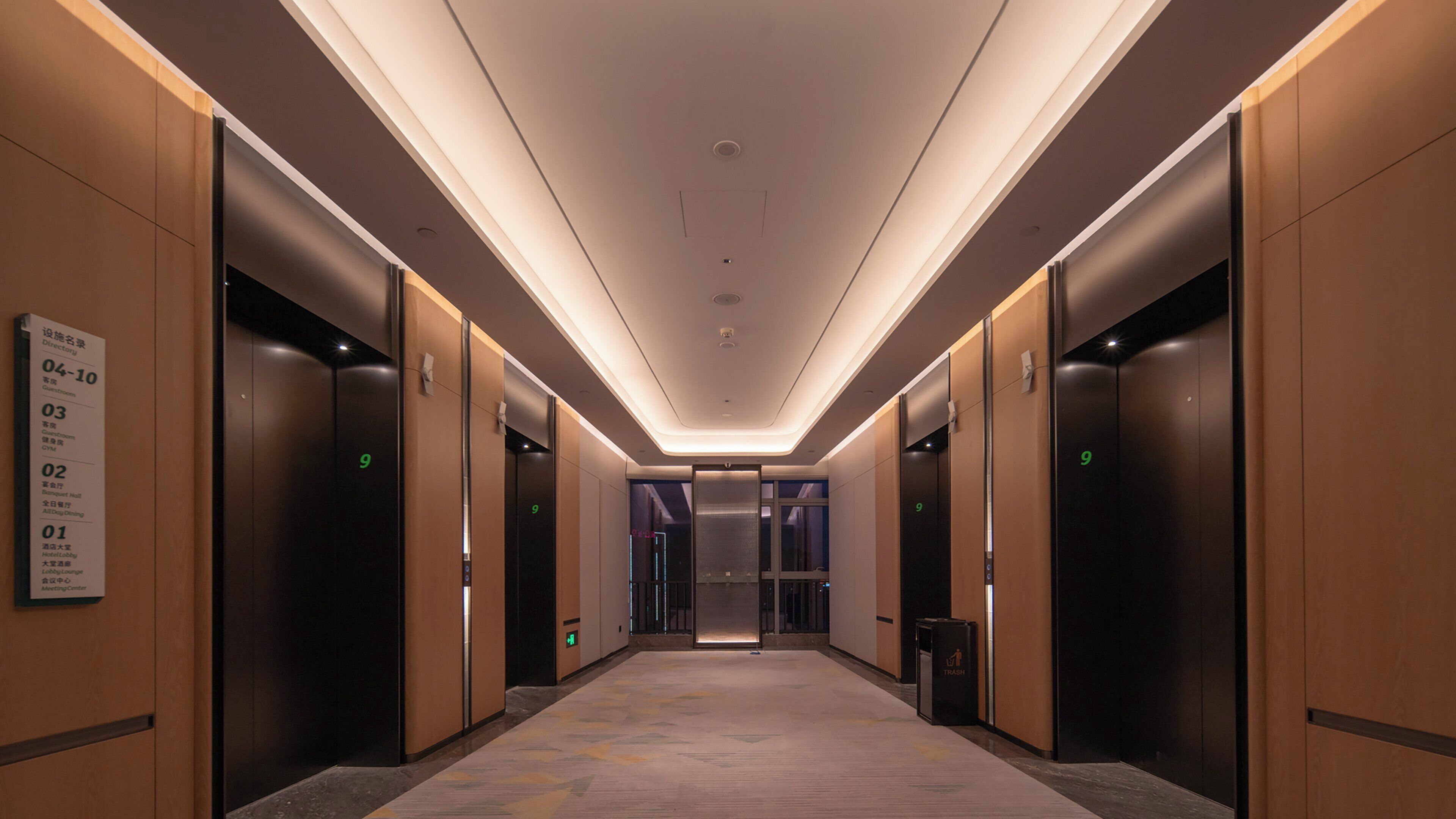 A modern hotel corridor with multiple elevators, a directory sign, and a trash bin.