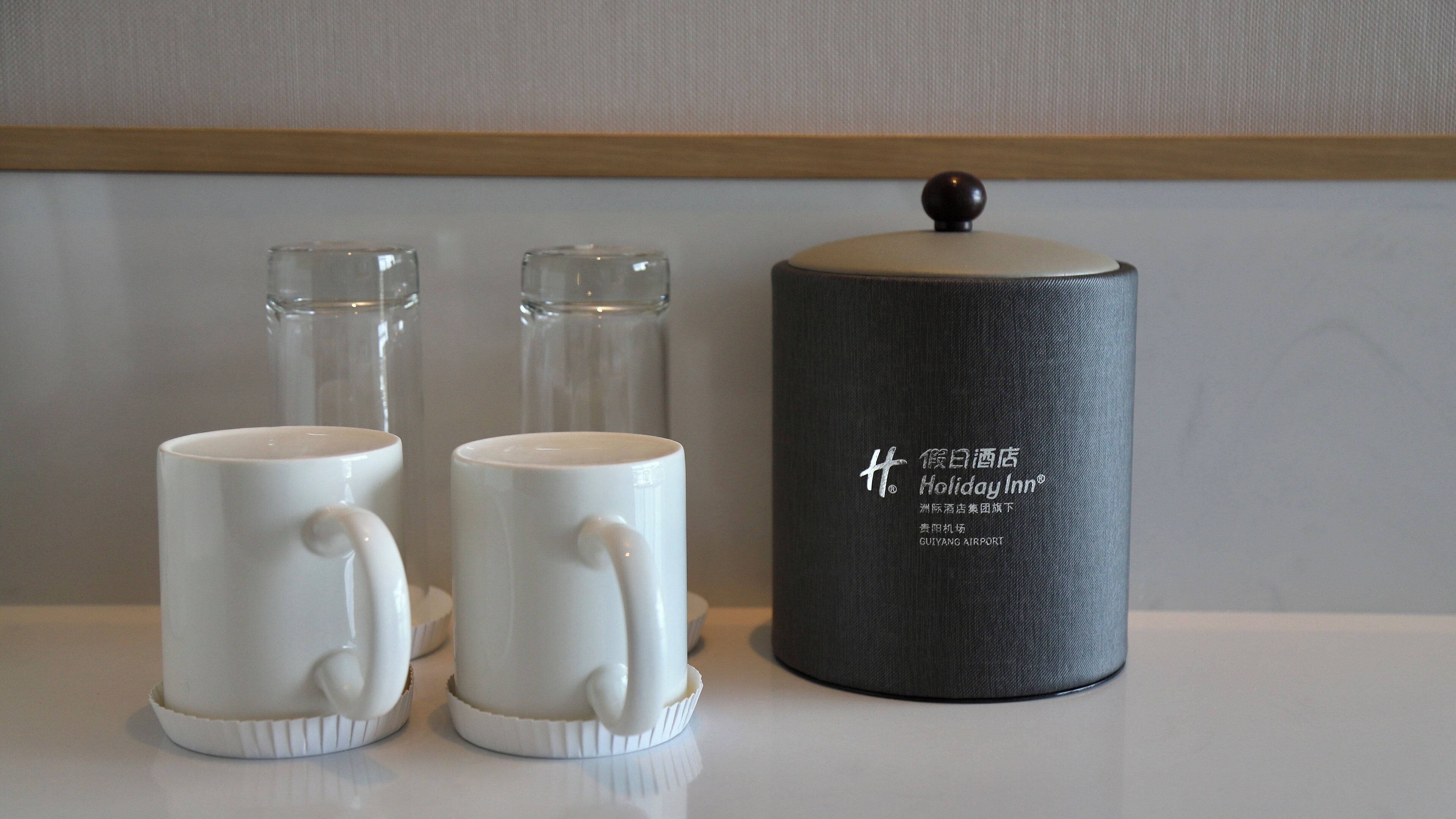 Two white ceramic mugs with handles, a glass container, and a black cylindrical object with text on it.