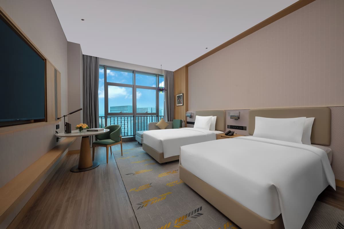 Une chambre d’hôtel avec deux lits, un bureau, une chaise, une grande fenêtre donnant sur un beau paysage et un balcon.