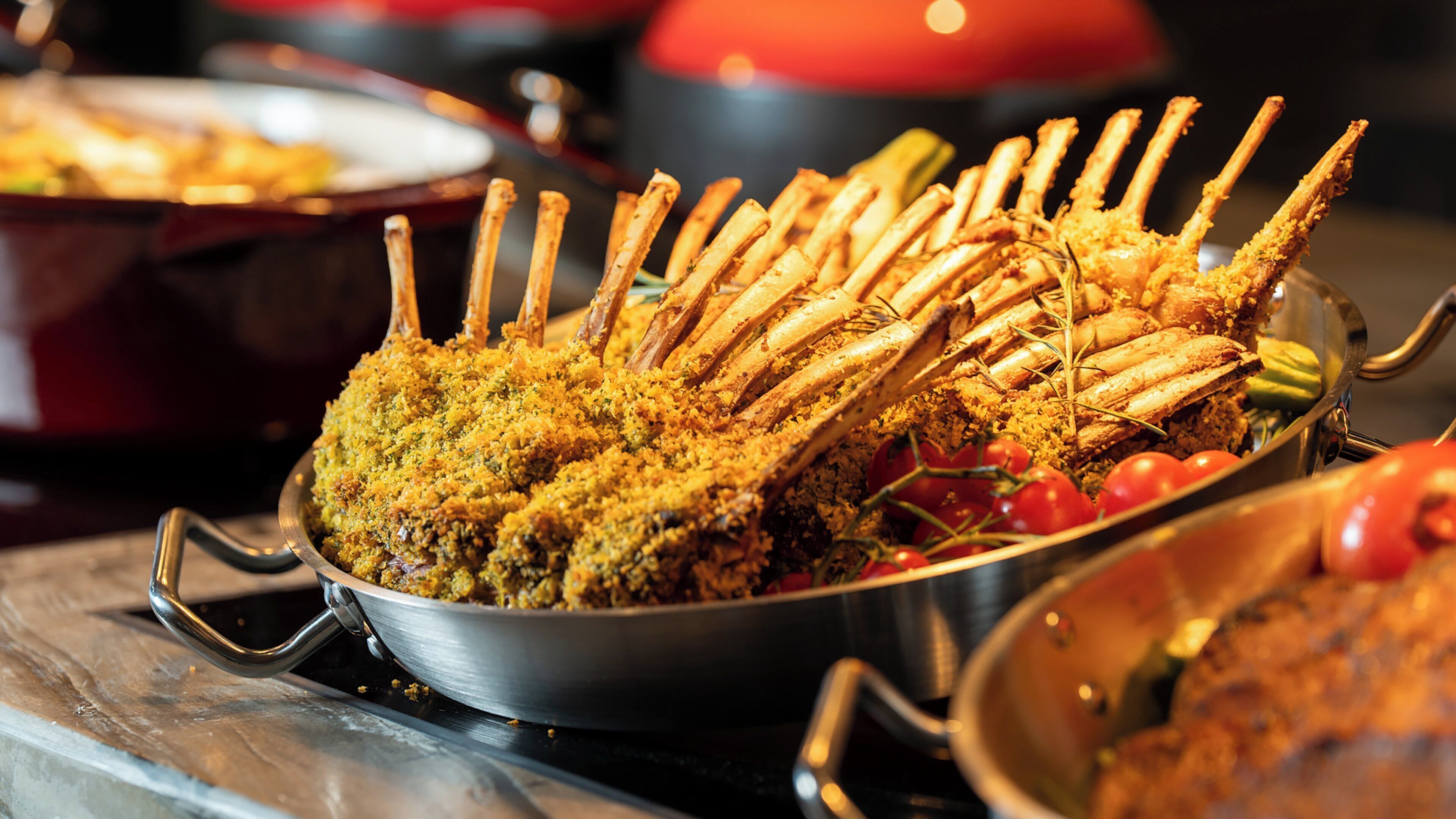 A roasted rack of lamb with a golden-brown crust, garnished with herbs and surrounded by a metal serving dish.