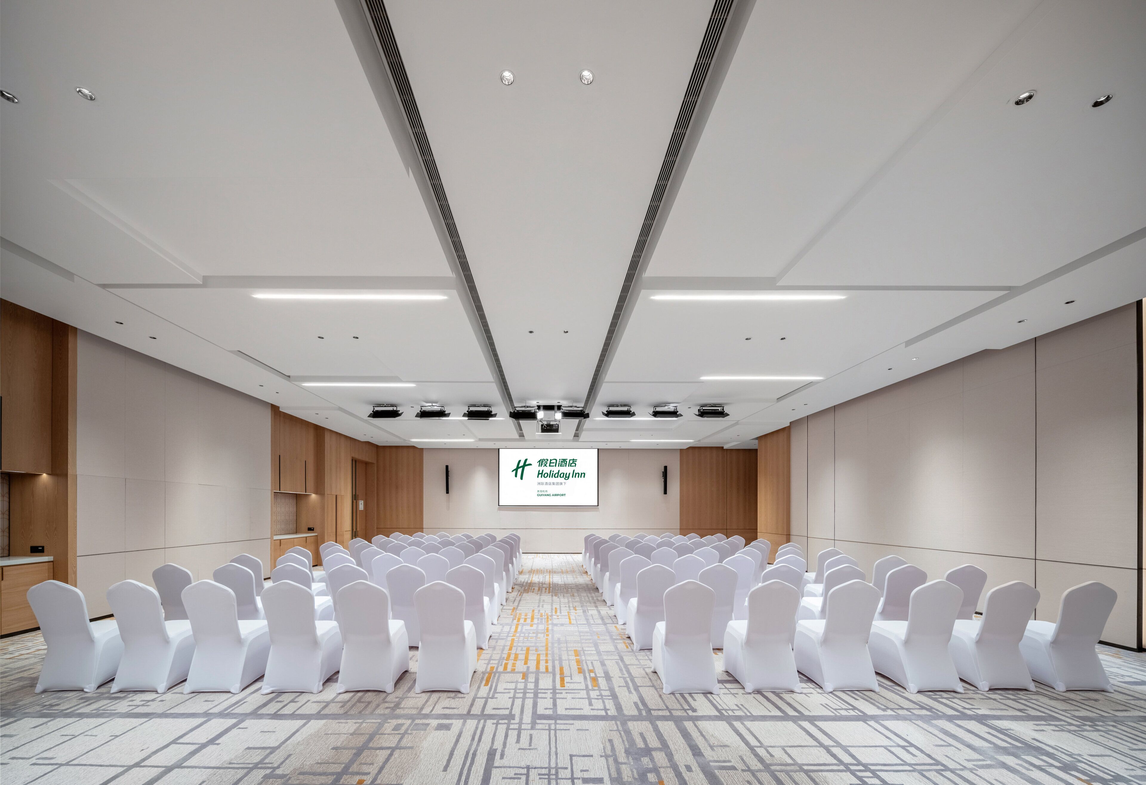 A conference room with rows of white chairs, a large screen at the front, and a carpeted floor.