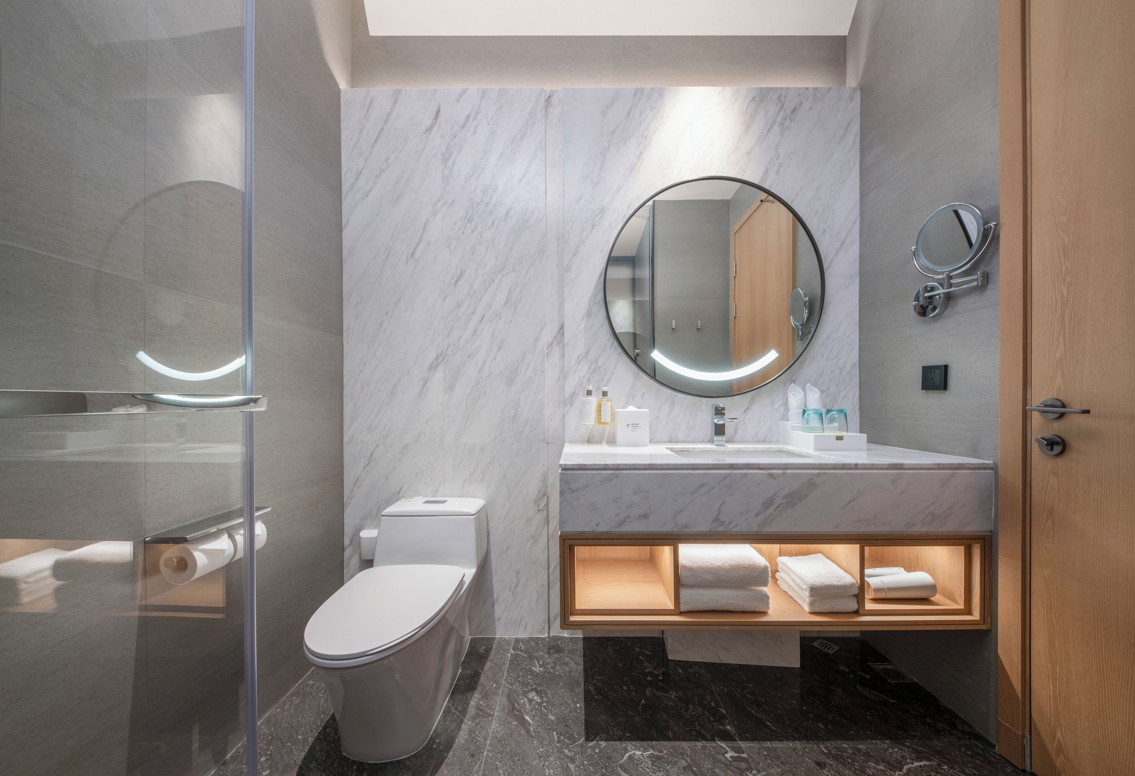 A modern bathroom with a glass shower enclosure, a toilet, a sink with a mirror, and a wooden vanity with towels.