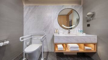 Un bagno moderno con una parete in marmo, uno specchio rotondo, un WC con maniglie di sostegno, un mobile lavabo con piano in marmo e uno scaffale con asciugamani.
